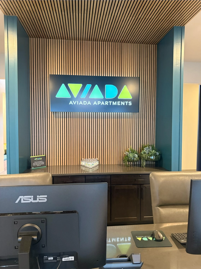  Backlit lobby sign for Aviada Apartments mounted on a wood slat wall behind a leasing office reception desk